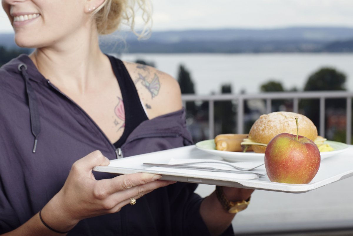 [Translate to Englisch:] Eine junge Frau hält ein Tablett auf dem ein Apfel, zwei Bockwürste, ein Käsebrötchen und Besteck ist.