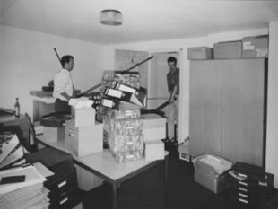 Room with packed tables and lots of boxes. Two people carry a table out of the door. 